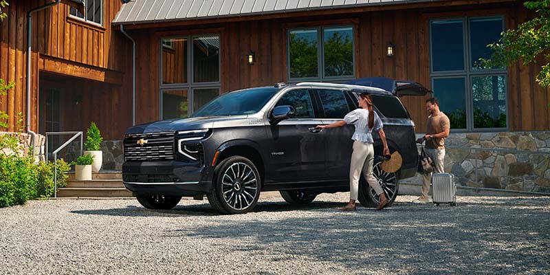 Black Chevrolet Tahoe parked in a gravel driveway as a couple loads luggage into the open trunk.
